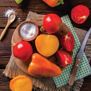 Red and orange peppers and tomatoes on a round wood slice with a green and white napkin and knife flat lay